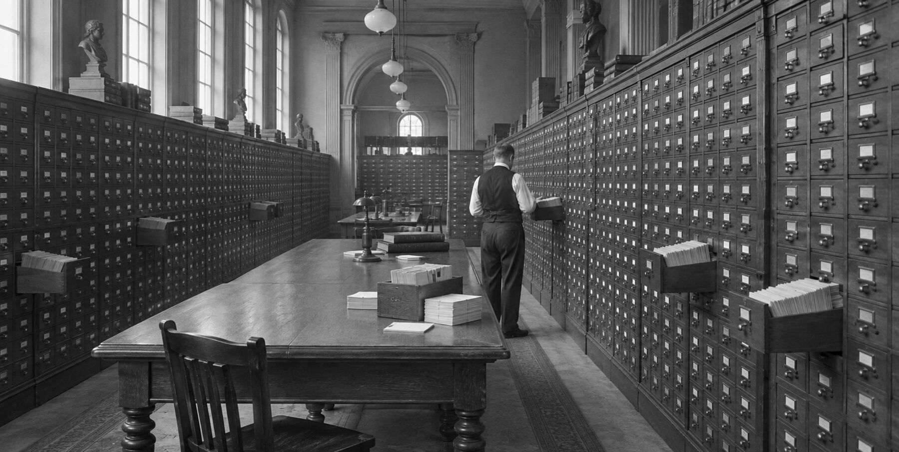 Sala de fichero de una biblioteca a fines del siglo XIX: paredes de cajoncitos de fichas y un archivista ordenando.